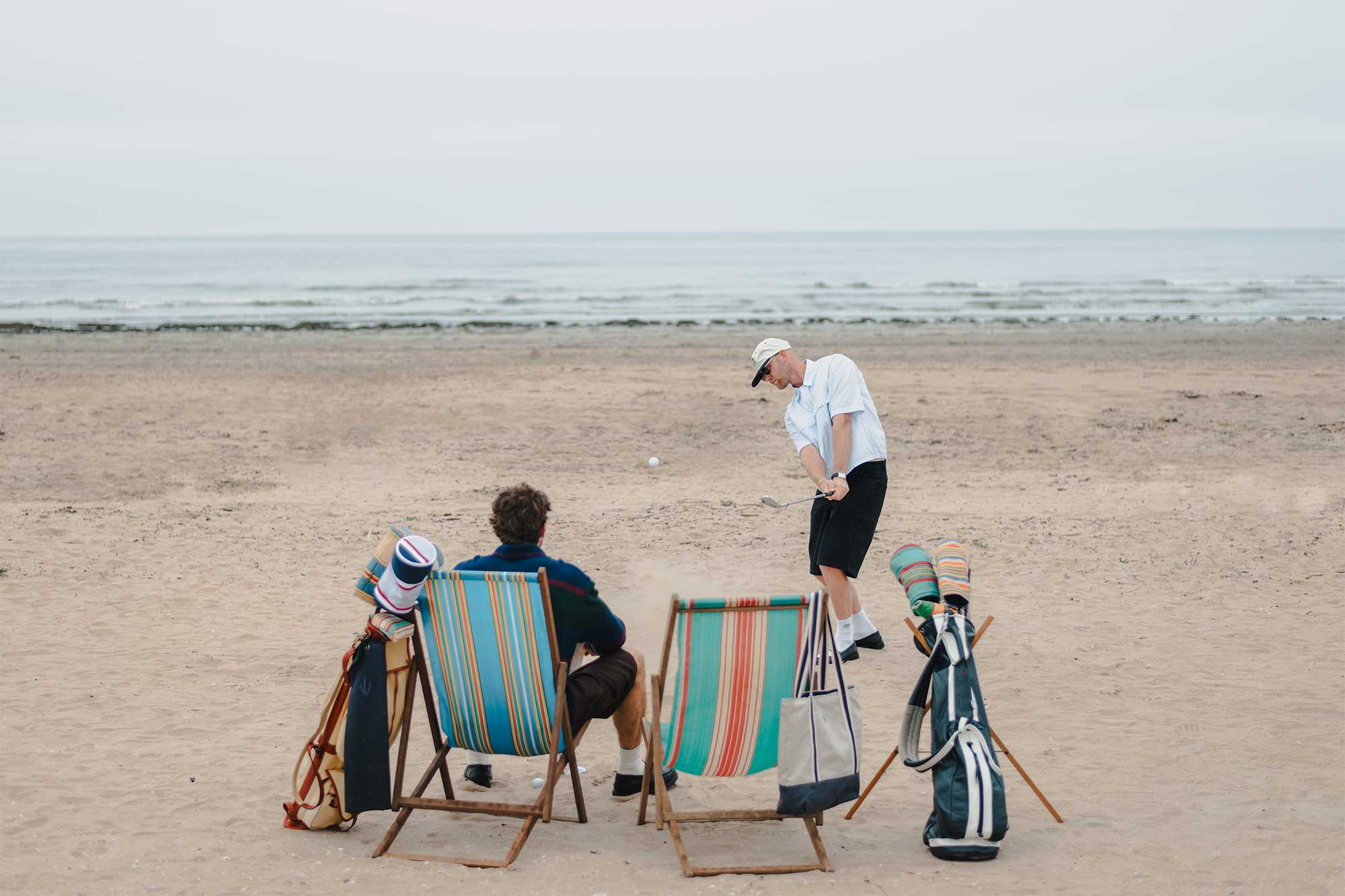 An image of two golfers playing golf on the beach at West Sands in St Andrews. One is taking small lob shot whilst the other sits on a vintage deck chair alongside a golf bag curated with Fyfe Golf's headcovers.