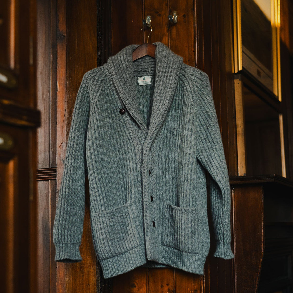 A ribbed knit shawl-collar cardigan sweater in a muted blue-gray color, hanging on a wooden hanger against a dark wood-paneled wall.