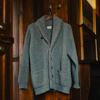 A ribbed knit shawl-collar cardigan sweater in a muted blue-gray color, hanging on a wooden hanger against a dark wood-paneled wall.
