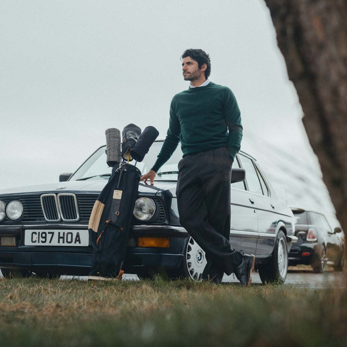 A man in a dark green sweater and black trousers stands beside a vintage BMW sedan outdoors, holding a golf bag loaded with clubs. The photo is taken from a low angle with a blurred tree trunk on the right and grass in the foreground.