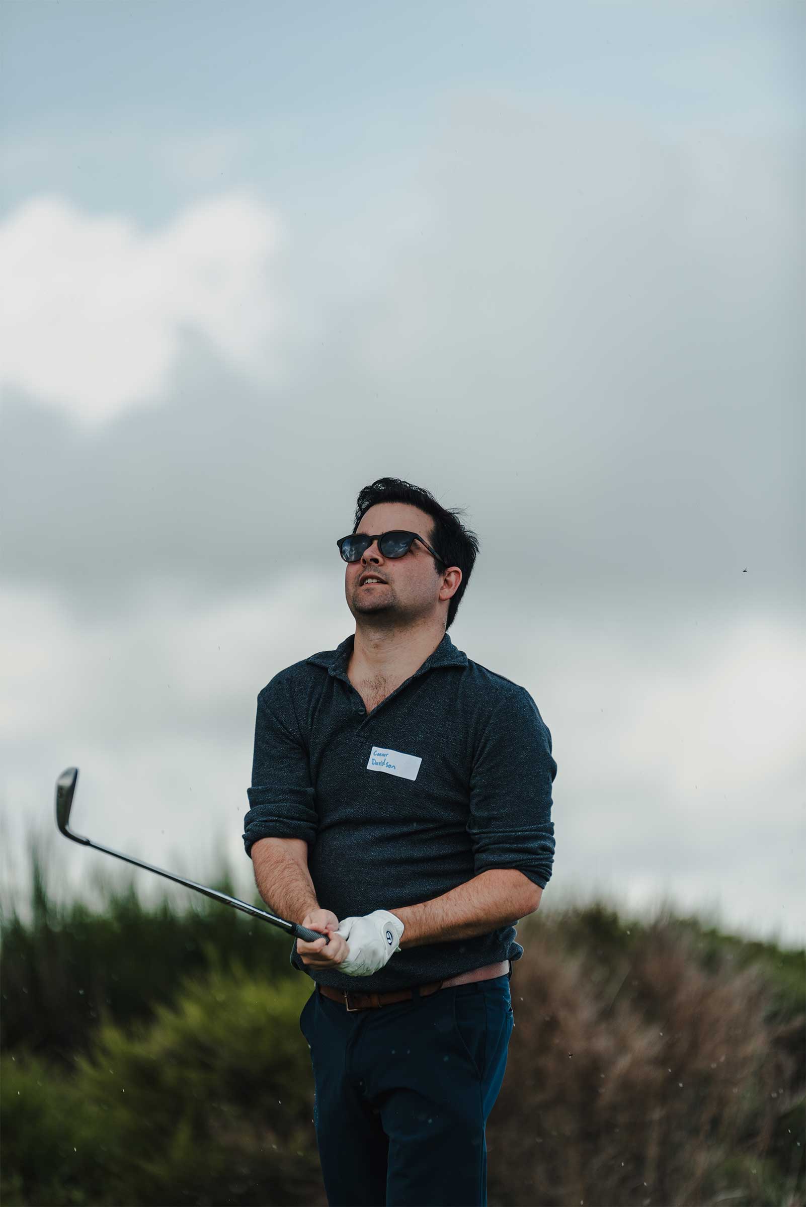 Golfer watching his shot just played at Spey Bay Golf course. Wearing sunglasses and a long sleeved polo with sleeves rolled up and holding the golf club post shot.