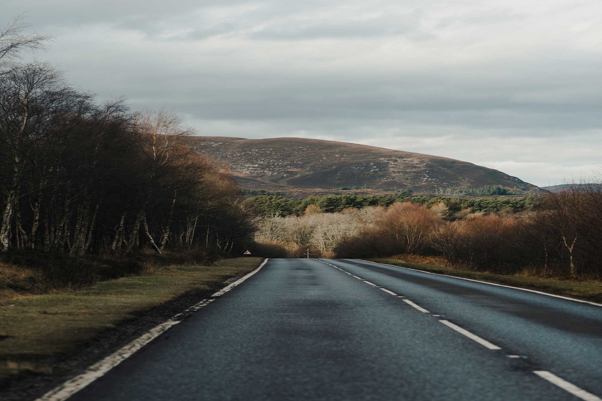 Scottish country road with mountain backdrop – A clear, winding country road in the Scottish Highlands, bordered by lush green fields and distant mountains, suggesting an adventurous journey ahead through the scenic and peaceful landscape.